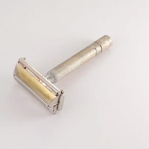 Peut inclure: Un rasoir de sécurité vintage avec un manche argenté texturé et une tête dorée. Le rasoir est conçu pour le rasage et présente un design classique et fonctionnel. Le rasoir est présenté sur fond blanc.