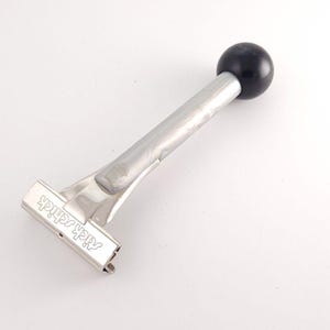Könnte beinhalten: Ein manueller Rasierer aus silberfarbenem Metall mit einem langen, zylindrischen Griff, der mit einem glatten, runden schwarzen Knopf versehen ist. Der Rasierkopf ist rechteckig und trägt am Ende des Griffs den Schriftzug "Schick" in Schreibschrift.