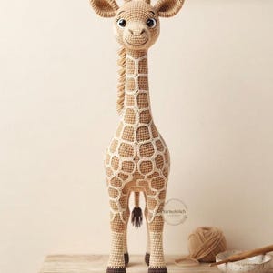 Könnte beinhalten: Ein gehäkeltes Giraffenspielzeug mit beigem Körper, weißen aufgestickten Flecken, dunkelbraunen Hufen und einer kleinen Mähne. Die Giraffe hat große dunkle Augen und ein aufgesticktes Lächeln. Ein kleines kreisförmiges Logo mit YarnoStitch ist sichtbar.