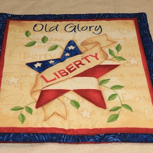 Może przedstawiać: Kwadratowy kawałek beżowego materiału z czerwoną, białą i niebieską gwiazdą oraz banerem z napisem "LIBERTY". Materiał ma vintage'owy wygląd z pismem odręcznym, które na górze mówi "Old Glory" i innymi słowami na całym materiale. Kwadrat jest obramowany czerwoną i niebieską tkaniną.