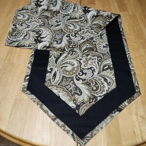 May include: A black and gold paisley table runner with a black border. The runner is folded in half and is lying on a wooden table.