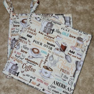 May include: Two square quilted pot holders with loop handles. The off-white fabric is printed with coffee-themed text and illustrations, including coffee cups, beans, and brewing equipment. The text includes "Coffee", "French Press", and "American".