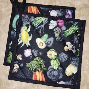 Op de afbeelding: Twee zwarte, gewatteerde pannenlappen met een kleurrijke groente print, waaronder wortelen, maïs en artisjokken. Elke pannenlap heeft een zwarte lus om op te hangen. De randen zijn afgewerkt met zwarte bies.