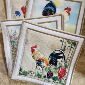 May include: Set of four square fabric placemats with a white border and a rooster design. The placemats feature a rooster with a red comb and wattles, a blue and black rooster, and a white rooster with a red comb and wattles. The placemats have a vintage, rustic look.