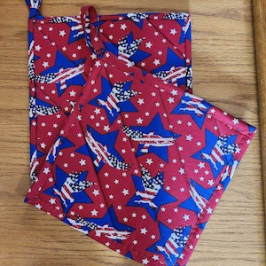 Könnte beinhalten: Zwei rote, gesteppte Topflappen mit einem blau-weißem amerikanischem Flaggenmuster. Die Topflappen haben eine Schlaufe zum Aufhängen.