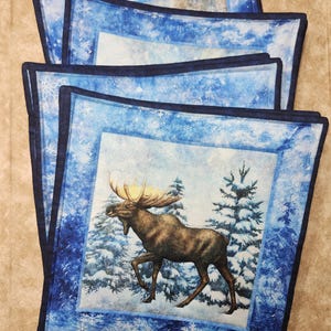 May include: Four blue and white throw pillows featuring a moose in a snowy forest scene. The pillows have a dark blue border and a white snowflake pattern.