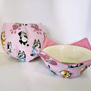 May include: Two pink bowl cozies with a cartoon dog print. One cozy is folded, and the other is shaped to hold a bowl, with pink flaps resembling ears. The print features various dog characters in different poses.