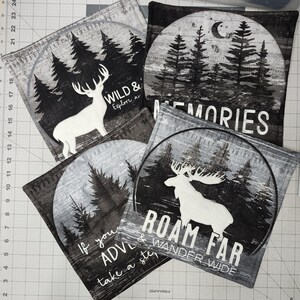 May include: A collection of square fabric coasters with nature-themed designs. Each coaster features a black and white illustration of trees, deer, or moose, with phrases like "WILD & Explore", "MEMORIES", and "ROAM FAR & WANDER WIDE".