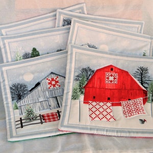 May include: Set of four red and white quilted fabric coasters featuring a winter scene with a red barn, a white fence, and a snowy landscape. The coasters have a blue and white border.