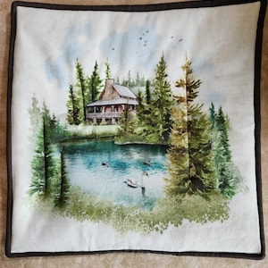 May include: Square cushion featuring a watercolour painting of a cabin by a lake. The scene includes evergreen trees, a lake with ducks and a swan, and a cabin with a stone chimney. The cushion has a dark brown border.