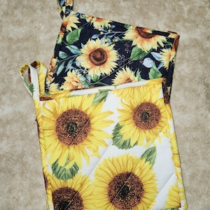 May include: Two quilted pot holders with sunflower patterns. One has a black background with yellow sunflowers and green leaves. The other has a white background with yellow sunflowers and green leaves. Both have a loop for hanging.