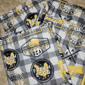 May include: A stack of quilted pot holders with a black and white checkered pattern. The pot holders feature yellow and black circular and rectangular designs with text that reads "Fresh Eggs", "Buy Fresh Grown", and "Farm Support Local".