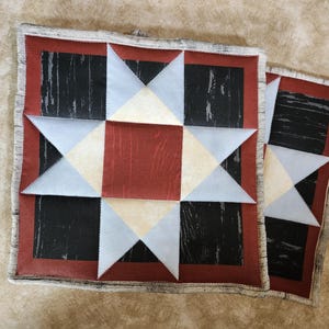 Puede incluir: Dos cuadrados de colcha de retazos rojos, blancos y azules con un patrón de veta de madera negro y blanco en el centro. Los cuadrados están cosidos juntos para crear un patrón de estrella.