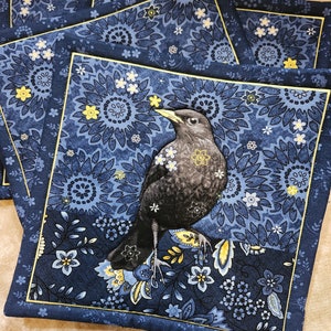 May include: Set of four blue fabric coasters with a yellow floral pattern and a black bird illustration. The coasters have a yellow border.