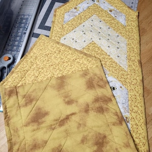 May include: A yellow and brown quilted table runner with a honeycomb pattern and a design of three white arrows. The runner is made of fabric with a subtle distressed look.