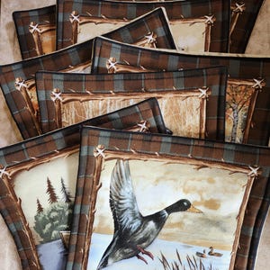 Op de afbeelding: Een stapel bruine en groene stoffen onderzetters met een ruitpatroon en een houten rand. De bovenste onderzetter heeft een schilderij van een eend in vlucht boven een meer.