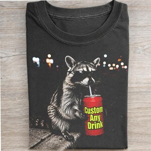 Pode incluir: Uma t-shirt cinzento-carvão com o gráfico de um guaxinim a usar óculos de sol pretos e a segurar uma lata de refrigerante vermelha com uma palhinha. A lata tem o texto amarelo "Custom Any Drink". Fundo com luzes da cidade desfocadas.