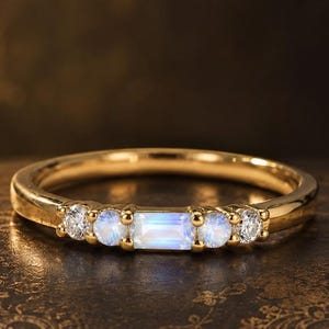 May include: A gold ring featuring a central baguette-cut moonstone flanked by two round moonstones and two round clear stones. The band is polished gold, and the stones are set in a horizontal row across the top of the ring's circular band.
