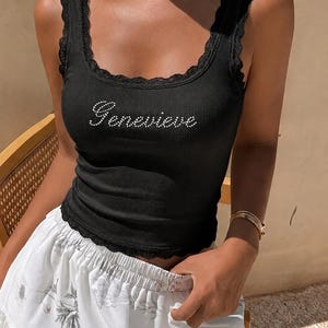 Puede incluir: Una camiseta de tirantes negra acanalada con escote redondo adornado con encaje negro y el nombre Genevieve escrito en pequeños brillantes blancos en el pecho. La prenda tiene una silueta ajustada con un dobladillo de encaje festoneado en la parte inferior.