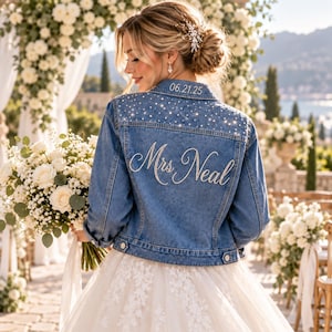 May include: A blue denim jacket featuring a collar embroidered with the date 06.21.25, shoulders embellished with scattered clear rhinestones, and the back decorated with the name Mrs. Neal in cursive script made of small white pearls.