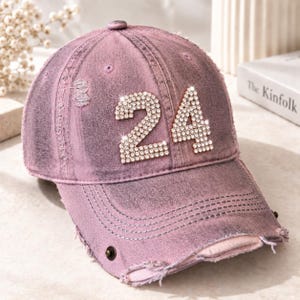 May include: A distressed mauve baseball cap featuring the number 24 on the front panel. The number is constructed from small, clear, reflective rhinestones. The cap has a worn-in appearance with frayed edges along the brim and crown panels.