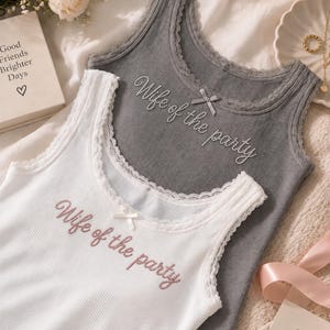 Op de afbeelding: Een wit geribbelde tanktop en een grijze geribbelde tanktop, beide voorzien van kanten afwerking langs de halslijn en armsgaten, een klein strikje in het midden en de tekst "Wife of the party" geborduurd in een sierlijk lettertype op de borst.