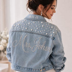 Puede incluir: Una chaqueta de mezclilla azul claro con adornos de perlas blancas en la parte superior de la espalda y los hombros. El texto "Mrs. Johnson" está escrito con pedrería plateada en el centro de la espalda y la fecha "06.21.25" en el cuello.