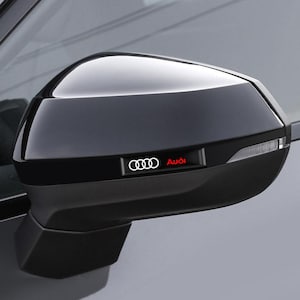 Peut inclure: Un rétroviseur extérieur automobile noir brillant doté d'un boîtier aérodynamique. Un petit emblème rectangulaire est intégré sur le côté, affichant le logo Audi à quatre anneaux blancs et le texte Audi en rouge. Un clignotant clair est inclus.