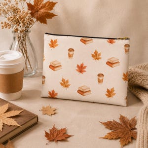 Peut inclure: Une pochette rectangulaire à fermeture éclair avec un motif répétitif de feuilles d'érable orange, de piles de livres et de tasses à café sur un fond crème. La pochette possède une fermeture éclair noire et une tirette en métal doré.