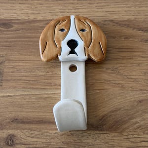 May include: A ceramic wall hook featuring a hand-painted beagle dog head at the top. The dog has tan ears, a white face, and a black nose. Below the dog head is a cream-coloured vertical stem with a circular mounting hole and a curved hook at the base.