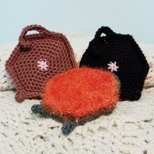 May include: Three crocheted dishcloths, one brown, one black, and one orange with a sparkly texture. Each dishcloth has a pink flower embroidered on it.