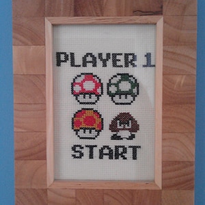 May include: Cross-stitch artwork in a wooden frame. The design features the words "PLAYER 1" and "START" above and below four mushroom graphics. The frame is made of light-colored wood.