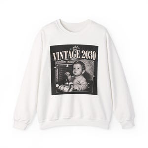 Könnte beinhalten: Ein weißes Langarm-Sweatshirt mit einer schwarzen quadratischen Grafik auf der Brust. Die Grafik zeigt ein Schwarz-Weiß-Foto eines Babys mit dem Text VINTAGE 2030 darüber und einem kleinen Kronensymbol, das oben mittig platziert ist.