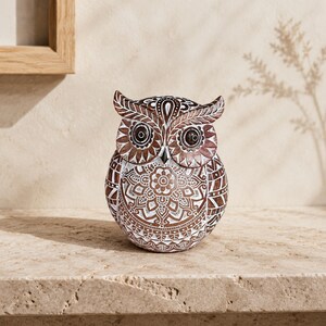 May include: A brown and white owl figurine featuring intricate mandala-style patterns etched across its surface. The owl has large, dark, circular eyes and pointed ear tufts, with a detailed floral design centered on its chest and geometric motifs.
