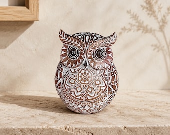 May include: A brown and white owl figurine featuring intricate mandala-style patterns etched across its surface. The owl has large, dark, circular eyes and pointed ear tufts, with a detailed floral design centered on its chest and geometric motifs.