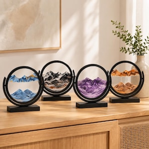May include: Four circular sand art displays with black frames and rectangular bases. Each piece contains colored sand forming mountain landscapes in blue, black and white, purple, and orange tones, encased between two clear glass panes.