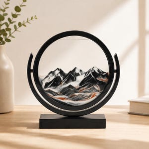 Black and white 3D moving sand art forming mountain scenery, modern round glass frame tabletop decor.