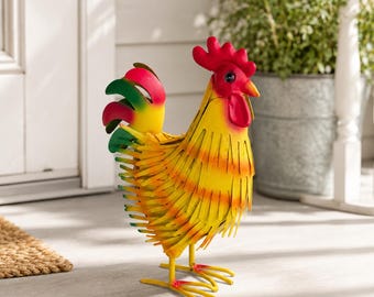 May include: A metal rooster sculpture featuring a bright yellow body with orange-tipped feathers, a vibrant red comb and wattle, and a tail with green, yellow, and red accents. The figure stands on two thin metal legs with pointed feet.