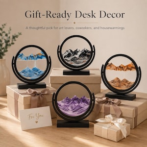 May include: Four circular sand art displays with black frames and rectangular bases, each containing layered sand forming mountain landscapes in blue, black and white, orange, and purple, arranged with gift boxes and a card reading For You.