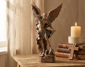 May include: A bronze-colored statue of an angel with large wings, wearing armor and holding a sword, standing over a figure. Beside the statue are three stacked vintage books, a small wooden cross, and a lit cream-colored pillar candle.