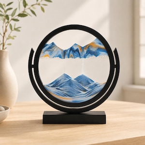 May include: A circular black frame holds a glass disc containing layers of blue, white, and gold sand. The sand is arranged to form mountain-like peaks and valleys within the clear glass, supported by a rectangular black base stand.