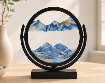 May include: A circular black frame holds a glass disc containing layers of blue, white, and gold sand. The sand is arranged to form mountain-like peaks and valleys within the clear glass, supported by a rectangular black base stand.
