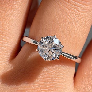 Op de afbeelding: Een solitaire ring met een ronde, briljant geslepen heldere edelsteen in een zilverkleurige metalen zetting met zes pootjes. De centrale steen heeft meerdere facetten die het licht reflecteren en de ringband heeft een gladde, gepolijste afwerking.