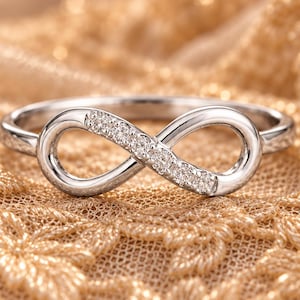 May include: A silver-toned ring featuring an infinity symbol as the centerpiece. One side of the infinity loop is embellished with a row of small, clear, sparkling stones set into the metal band, creating a polished and reflective appearance.