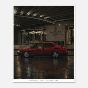 May include: A red 1987 Saab 900 Turbo sedan parked on a wet surface in front of the Grand Prince Hotel Hiroshima. The car features three-spoke wheels and a dark lower trim. The text 1987 SAAB 900 TURBO - HIROSHIMA, JAPAN is printed below.