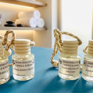 Può includere: Quattro piccoli flaconi in vetro trasparente per profumatori auto con tappi in legno chiaro e cordini per appenderli. Ogni etichetta bianca riporta il testo SAAM SERENITY CANDLE & HOME e le fragranze Strawberry Guava, Himalayan Orchid, Pumpkin Pecan Delight e Jasmine Bloom.