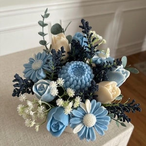 Può includere: Una composizione floreale con fiori scolpiti color azzurro e crema, tra cui margherite, rose e tulipani. Il bouquet è arricchito da rametti blu scuro, piccoli fiori bianchi di riempimento e foglie verdi in stile eucalipto.