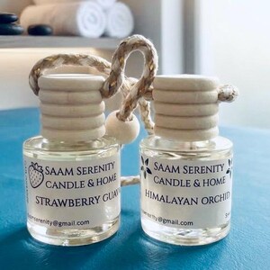 Puede incluir: Dos frascos pequeños de vidrio transparente con tapones de madera clara y cuerdas para colgar. Las etiquetas dicen Saam Serenity Candle & Home, con Strawberry Guava y Himalayan Orchid, incluyendo la dirección saamserenity@gmail.com.