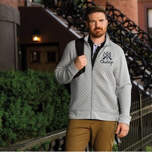 May include: A gray quilted jacket with a zipper closure. The jacket has a black backpack strap and a navy blue embroidered logo that says "Lake Chatuge" with crossed oars.