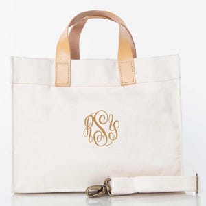 May include: Off-white canvas tote bag with a gold monogram. Features light brown leather handles and a detachable shoulder strap. The bag has a rectangular shape and a metal clasp.